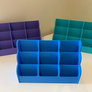 May include: Three desktop organizers with six compartments each. The organizers are made of plastic and are blue, teal, and purple.