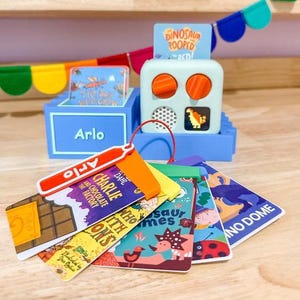 May include: A blue toy cash register with a red handle and a variety of colorful book-shaped tags. The tags feature titles like "Charlie and the Chocolate Factory" and "Dinosaur Homes".