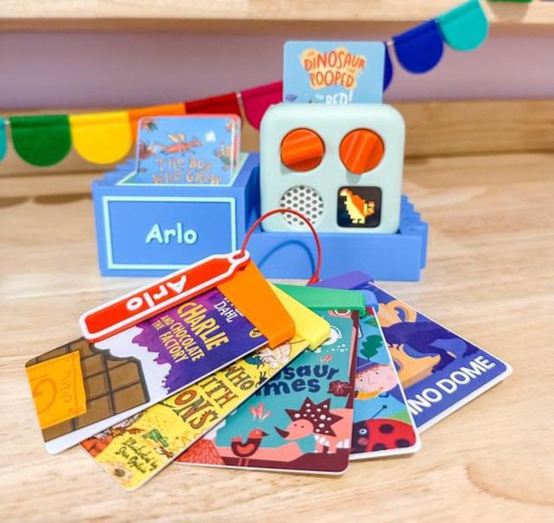 May include: A blue toy cash register with a red handle and a variety of colorful book-shaped tags. The tags feature titles like "Charlie and the Chocolate Factory" and "Dinosaur Homes".