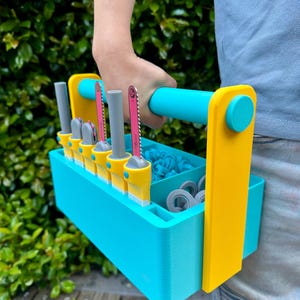May include: A vibrant blue and yellow toolbox with a handle, containing small saws and other tools. The toolbox is held by a person. The saws have pink blades and grey handles.