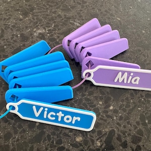 May include: Two sets of colorful plastic name tags with white lettering. One set is blue and says "Victor" and the other set is purple and says "Mia".