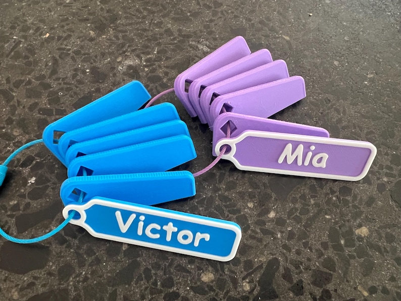 May include: Two sets of colorful plastic name tags with white lettering. One set is blue and says "Victor" and the other set is purple and says "Mia".