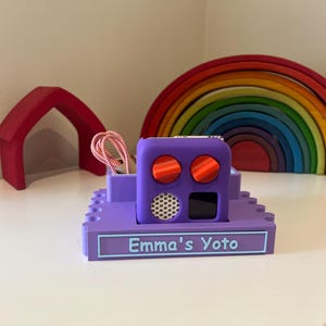 May include: Purple toy organizer with a purple and red Yoto player, a red and white cord, and a label that says "Emma's Yoto".