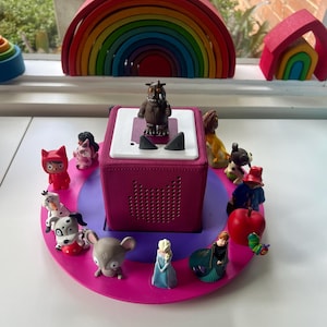 Może przedstawiać: Różowa i fioletowa okrągła platforma z różnymi figurkami, w tym brązowym stworzeniem, otoczona przez inne kolorowe postacie. Różowa kostka z białym wierzchem znajduje się na środku. Drewniana tęcza i inne zabawki znajdują się w tle.