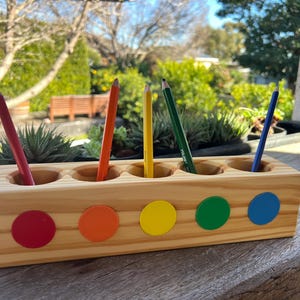 Pode incluir: Porta-lápis de madeira com cinco orifícios, cada um com um lápis de cor. O suporte tem cinco círculos coloridos: vermelho, laranja, amarelo, verde e azul. Os lápis são vermelhos, laranja, amarelos, verdes e azuis. O suporte é de madeira clara.
