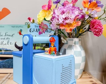 Puede incluir: Un reproductor de audio azul claro con un asa azul y el texto "Travelling Tales". Una pequeña figura de dinosaurio naranja está encima. Un jarrón de flores coloridas está en el fondo.