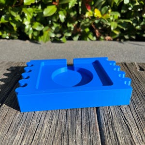 May include: A bright blue ashtray with a unique design. The ashtray has a circular indentation in the center and a rectangular shape with notched edges. The ashtray is made of a solid material and sits on a wooden surface.