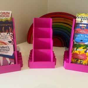 May include: Three pink plastic bookends with different children's books displayed. The bookends have three shelves each. The books include "Robin Hood", "The Secret Garden", "Alice's Adventures in Wonderland", "The Famous Five", "Home Flamingo", "Row Your Boot and other Pre-School Songs", and "Flamingo".
