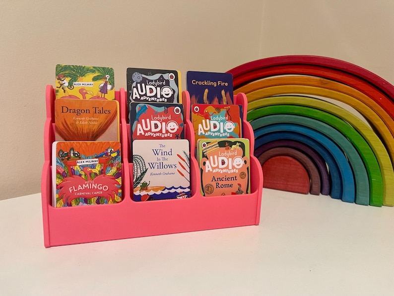 May include: A pink stand with eight slots holds a variety of children's books with colorful covers. The books feature titles like "Dragon Tales", "The Wind in the Willows", and "Ancient Rome". A rainbow-colored wooden toy sits in the background.
