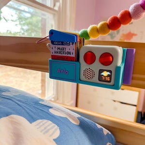 May include: A blue and white bedside alarm clock with a digital display showing 7:16. The clock is mounted on a wooden bunk bed with a blue and white patterned pillow. The clock has two red buttons and a small purple compartment on the side. A rainbow coloured beaded garland hangs above the clock.