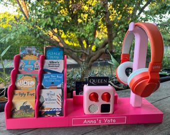 May include: Pink Yoto player with a pink headphone stand and a variety of children's books. The player has a screen that displays "QUEEN" and two red buttons. The headphone stand has the word "yoto" printed on it. The stand is labeled "Anna's Yoto".