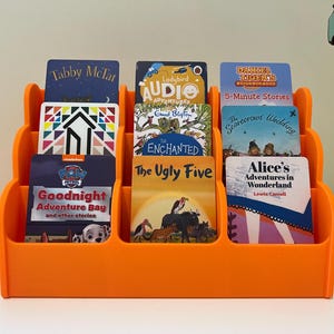 May include: Orange plastic book holder with 8 slots for books. The book titles include "Tabby McTat", "Ladybird Audio Adventures", "Goodnight Adventure Bay", "The Ugly Five", "Alice's Adventures in Wonderland", "The Scarecrows Wedding", "Enchanted", and "Daniel Tiger's 5-Minute Stories".