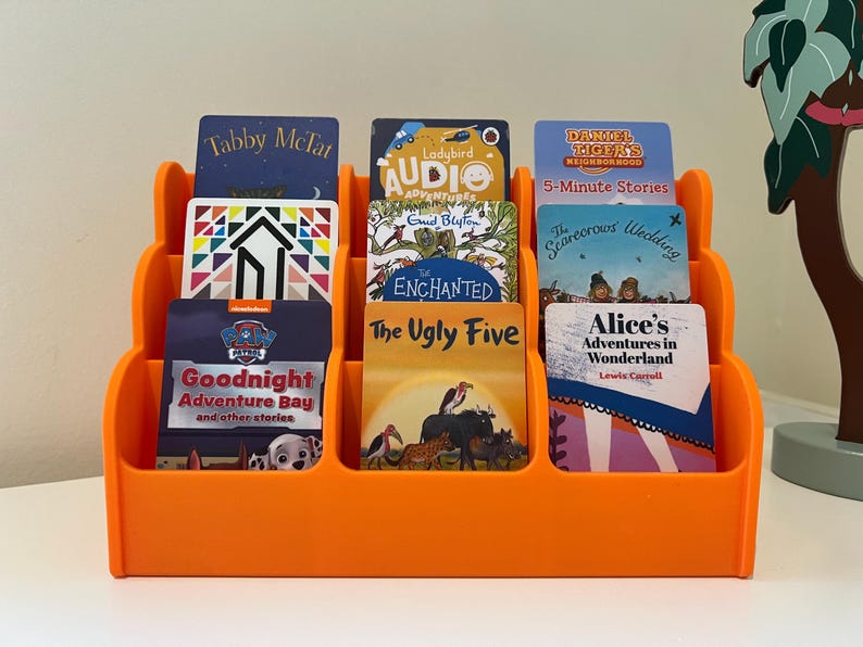 May include: Orange plastic book holder with 8 slots for books. The book titles include "Tabby McTat", "Ladybird Audio Adventures", "Goodnight Adventure Bay", "The Ugly Five", "Alice's Adventures in Wonderland", "The Scarecrows Wedding", "Enchanted", and "Daniel Tiger's 5-Minute Stories".