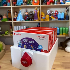 Puede incluir: Un contenedor de almacenamiento de plástico blanco que contiene varias cajas de juego rojas Tonies ToniePlay. La caja frontal presenta un gráfico morado con el texto The Quizzles y 500 questions. Un gran botón circular rojo con la palabra Play está fijado al frente.