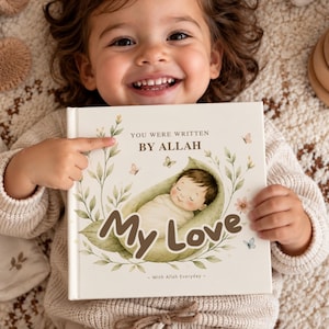 Peut inclure: Un livre à couverture rigide carré avec une couverture crème présentant une illustration à l'aquarelle d'un bébé emmailloté dans une feuille verte. Le texte indique "YOU WERE WRITTEN BY ALLAH My Love - With Allah Everyday -" entouré de feuillage vert et de papillons.
