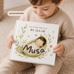 Peut inclure: Un livre carré à couverture rigide avec une couverture blanche portant le texte 'YOU WERE WRITTEN BY ALLAH' et le nom 'Musa' en grandes lettres marron. L'illustration montre un bébé enveloppé dans une feuille verte, entouré de feuillage.