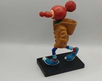 May include: A whimsical figurine of a character in a tan coat, blue pants, and blue shoes with white tips. The figure has a red head, nose, and two red pom-poms. It is posed as if walking on a black rectangular base.