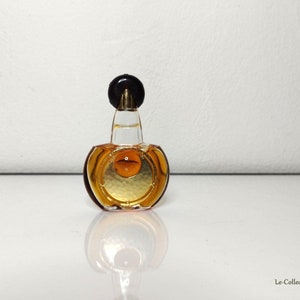 May include: A small, clear glass bottle of perfume with a gold-colored liquid and a black cap. The bottle has a round, textured design.