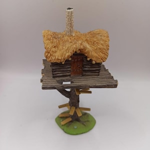 Op de afbeelding: Een miniatuur houten boomhut met een rieten dak en een kleine deur. De boomhut is gebouwd op een houten platform met een groene basis.