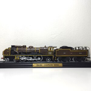 May include: A brown and black model train engine with yellow accents on a black display base. The base has a gold plaque that reads "PACIFIC CHAPELON NORD".