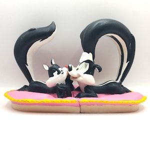 Puede incluir: Una figurita de pareja de mofetas de dibujos animados en blanco y negro, con la mofeta hembra acostada sobre una almohada rosa y la mofeta macho de pie sobre una almohada rosa, con las cabezas tocándose.