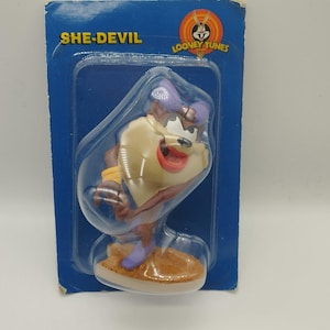 Peut inclure: Une figurine en plastique de Taz des Looney Tunes, le personnage est vêtu d'une tenue violette et jaune, la figurine est sur une base brune et est emballée dans un emballage en carton bleu et blanc avec les mots "SHE-DEVIL" et "LOONEY TUNES" imprimés dessus.