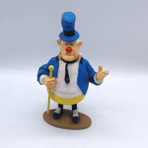 Peut inclure: Figurine de personnage de dessin animé avec un chapeau haut-de-forme et une veste bleus, un gilet jaune. Le personnage a un nez rouge, une cravate noire et tient une canne jaune. Il se tient sur une base ovale marron.