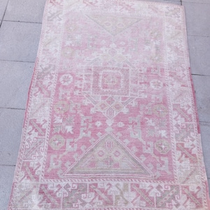 May include: A rectangular, vintage-style rug with a faded pink and off-white color palette. The rug features a central geometric design with a border of repeating patterns. The rug's distressed appearance suggests it is an antique or a reproduction.