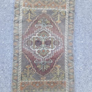 May include: A rectangular woven rug featuring a central diamond medallion in shades of muted red, brown, and beige. The rug displays a geometric pattern with stylized floral motifs and a decorative border with repeating orange and green elements.