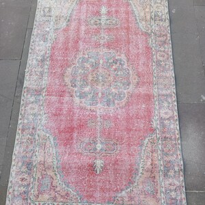 May include: A rectangular, distressed rug with a central red field and intricate border designs. The rug features a faded, vintage aesthetic with a mix of red, blue, and beige tones. The pattern includes floral and geometric motifs.