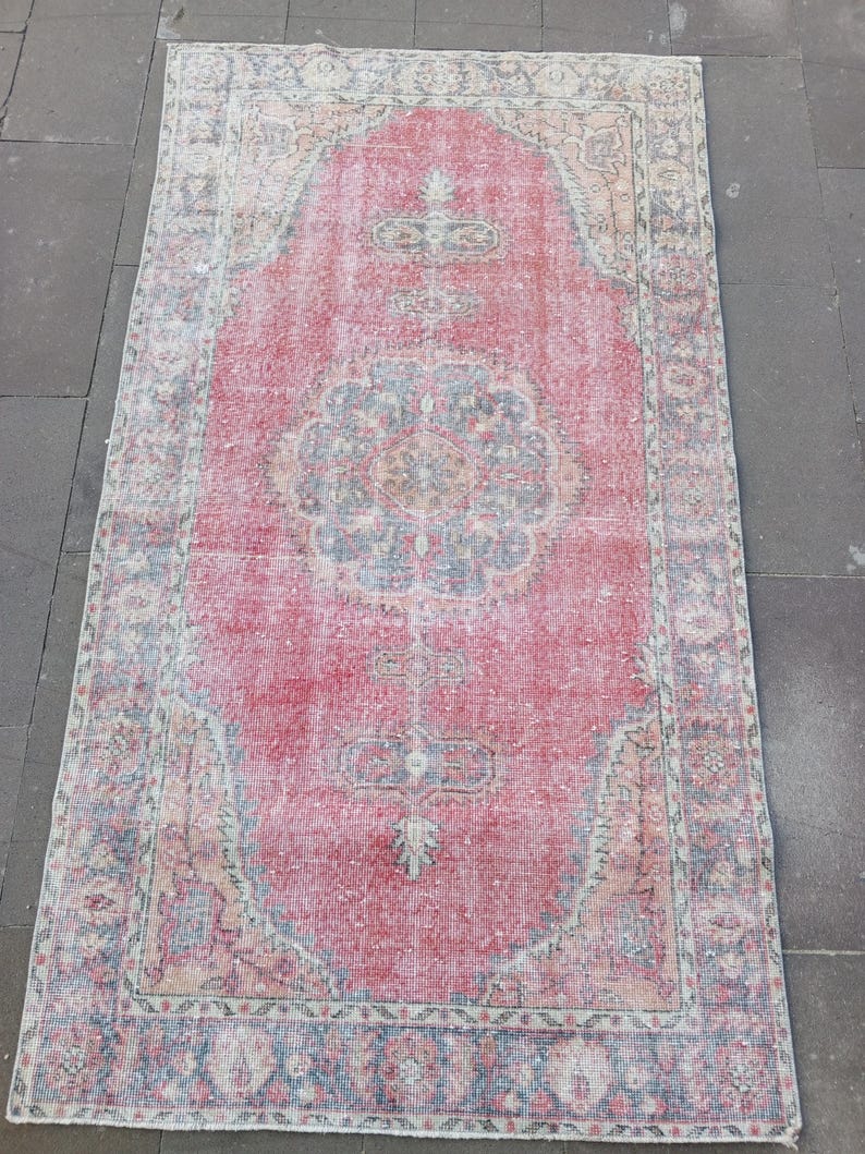 May include: A rectangular, distressed rug with a central red field and intricate border designs. The rug features a faded, vintage aesthetic with a mix of red, blue, and beige tones. The pattern includes floral and geometric motifs.