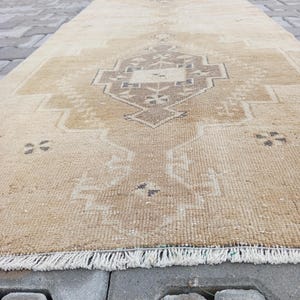 Może przedstawiać: Beżowy vintage dywan z geometrycznym wzorem w ciemniejszym odcieniu beżu. Dywan ma frędzlową krawędź i leży na szarej betonowej powierzchni.