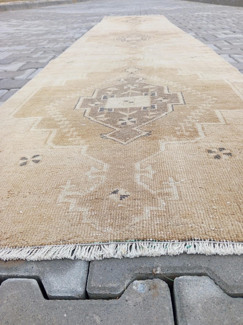 Może przedstawiać: Beżowy vintage dywan z geometrycznym wzorem w ciemniejszym odcieniu beżu. Dywan ma frędzlową krawędź i leży na szarej betonowej powierzchni.