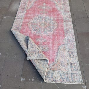 May include: A rectangular, distressed-look rug with a central medallion design. The rug features a faded red field with a detailed border and accents in blue, beige, and cream. The corner is folded over, revealing the underside.