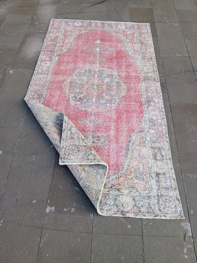 May include: A rectangular, distressed-look rug with a central medallion design. The rug features a faded red field with a detailed border and accents in blue, beige, and cream. The corner is folded over, revealing the underside.