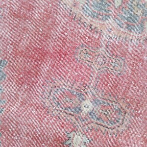 May include: Close-up of a distressed, vintage rug with a red and faded color palette. The rug features intricate geometric patterns in shades of blue, beige, and white, creating a textured, antique appearance. The rug's design suggests a traditional, oriental style.