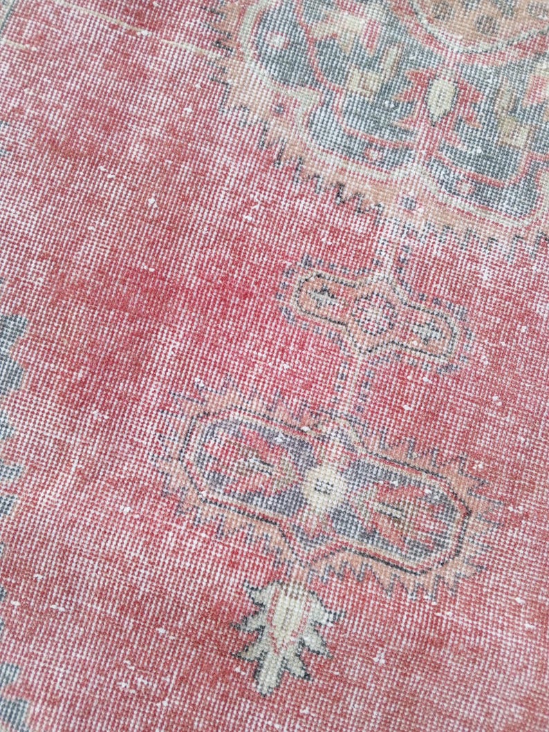 May include: Close-up of a distressed, vintage rug with a red and faded color palette. The rug features intricate geometric patterns in shades of blue, beige, and white, creating a textured, antique appearance. The rug's design suggests a traditional, oriental style.
