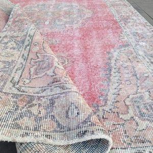 May include: A distressed, rectangular rug with a faded red central field and intricate border designs in muted tones of beige, blue, and brown. The rug is partially rolled, revealing its layered construction and aged appearance.