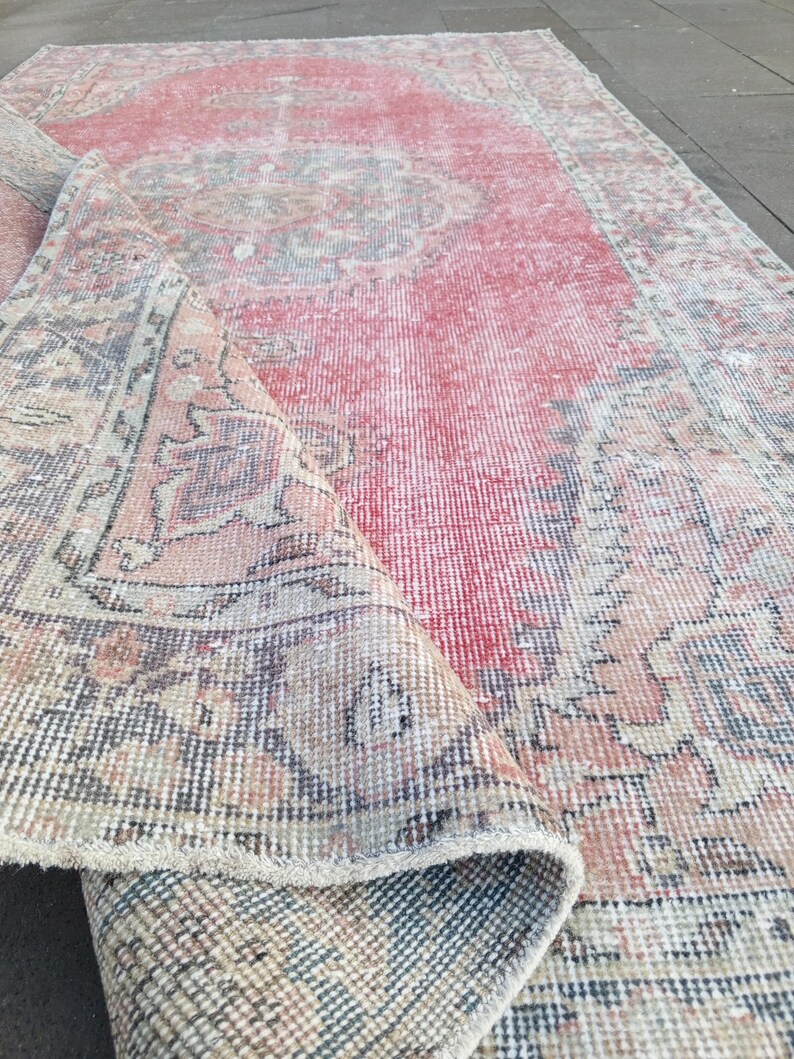May include: A distressed, rectangular rug with a faded red central field and intricate border designs in muted tones of beige, blue, and brown. The rug is partially rolled, revealing its layered construction and aged appearance.
