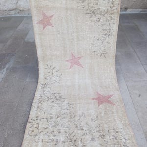 May include: Beige and faded red star patterned rug. The rug has three faded red stars and a floral design. The rug is rectangular and has a distressed, vintage look. The rug is being held up by a person.