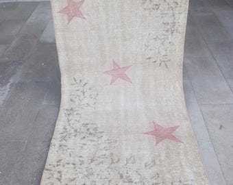 Puede incluir: Alfombra beige con estrellas rojas descoloridas. La alfombra presenta tres estrellas rojas descoloridas y un diseño floral. La alfombra rectangular tiene un aspecto vintage y desgastado. Una persona sostiene la alfombra.