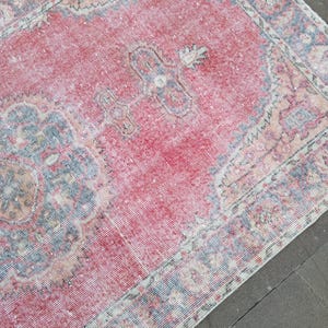 May include: A distressed, vintage rug with a faded red central field and intricate border designs in shades of blue, beige, and white. The rug has a worn, textured appearance, suggesting age and use. The overall aesthetic is traditional and antique.