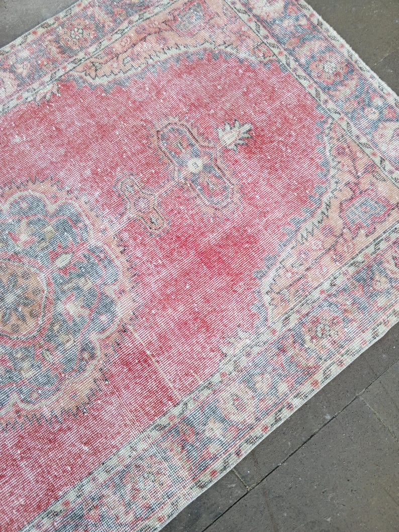 May include: A distressed, vintage rug with a faded red central field and intricate border designs in shades of blue, beige, and white. The rug has a worn, textured appearance, suggesting age and use. The overall aesthetic is traditional and antique.