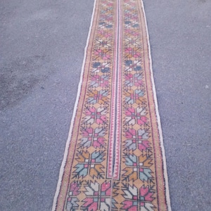 Pode incluir: Um tapete longo e estreito com um padrão geométrico em tons de rosa, azul, amarelo e marrom. O tapete tem franjas brancas nas bordas.