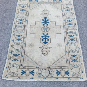 May include: A vintage Turkish rug with a cream background and a blue and brown geometric pattern. The rug has a fringed edge.