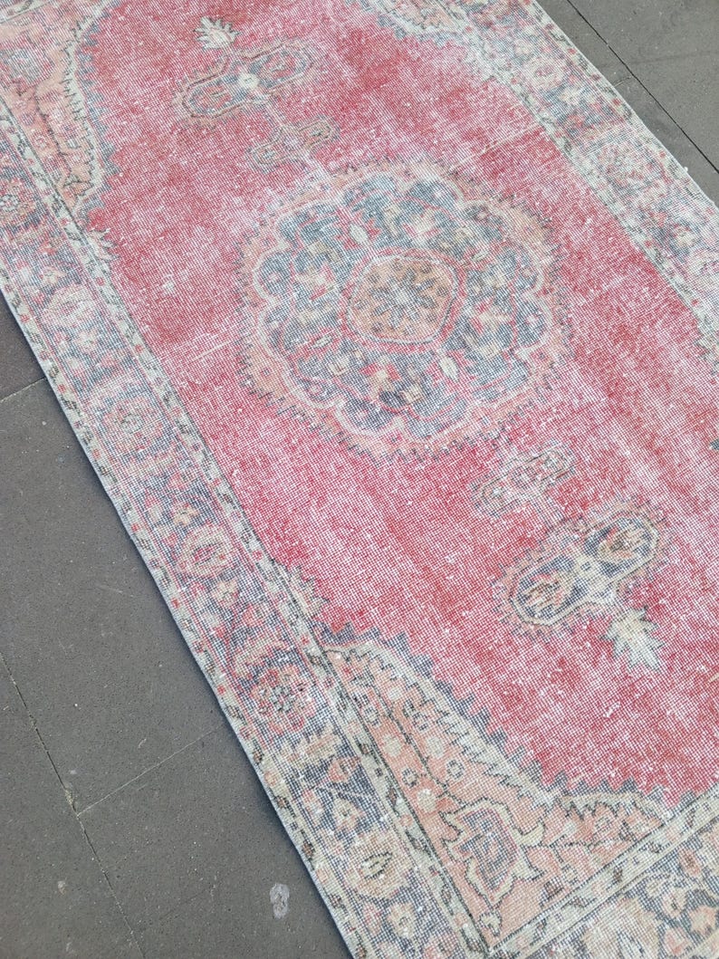 May include: A rectangular, distressed rug with a faded red central field and intricate floral patterns in blue, beige, and orange. The rug has a detailed border and a vintage aesthetic, suitable for home decor.