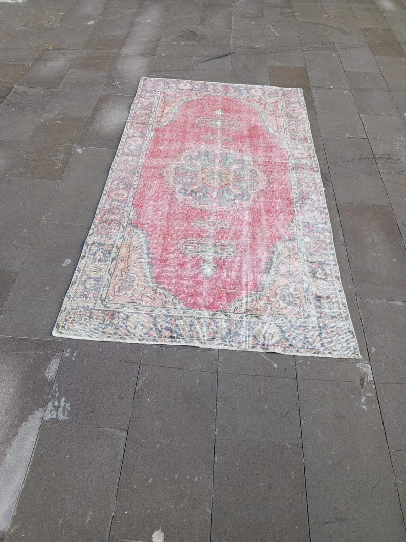 May include: A rectangular, distressed rug with a faded red central field and intricate floral patterns in shades of blue, beige, and cream. The rug's border features a similar design, creating a cohesive and vintage aesthetic. The rug is placed on a dark gray stone surface.