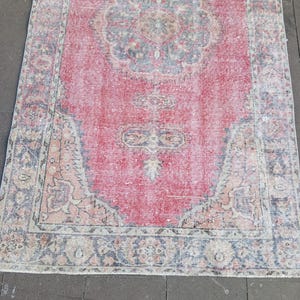 May include: A rectangular, distressed-look rug with a central medallion design. The rug features a faded red field with a detailed floral pattern in shades of blue, beige, and peach. The border is a darker blue with similar floral motifs.