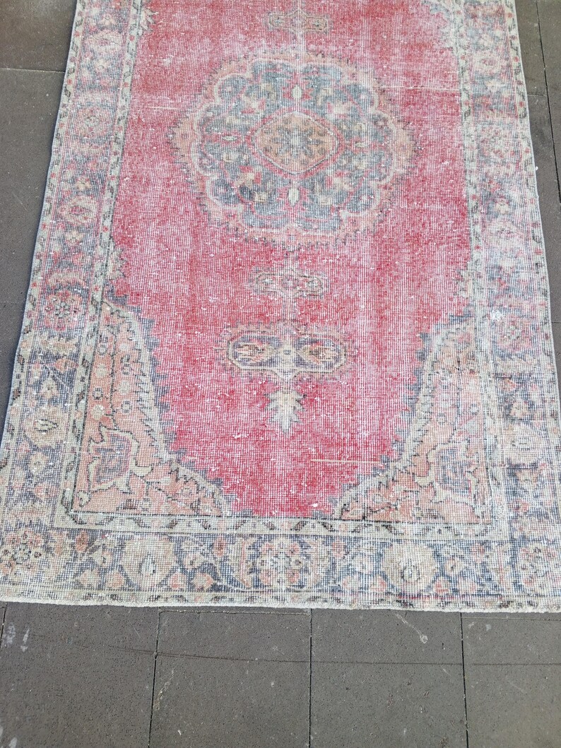 May include: A rectangular, distressed-look rug with a central medallion design. The rug features a faded red field with a detailed floral pattern in shades of blue, beige, and peach. The border is a darker blue with similar floral motifs.