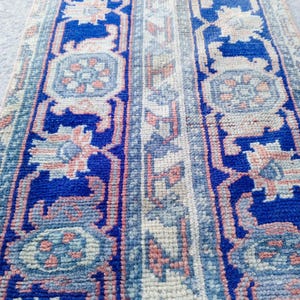 May include: A rectangular rug with a detailed pattern. The rug features a deep blue border with floral and geometric designs in shades of pink, beige, and light blue. The central section has a lighter, neutral color with similar patterns.